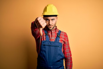 Young builder man wearing construction uniform and safety helmet over yellow isolated background looking unhappy and angry showing rejection and negative with thumbs down gesture. Bad expression.