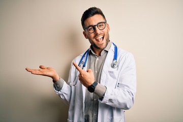 Young doctor man wearing glasses, medical white robe and stethoscope over isolated background amazed and smiling to the camera while presenting with hand and pointing with finger.
