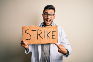 Young handsome doctor man protesting holding cardboard with strike message very happy pointing with hand and finger