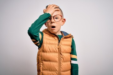 Young little caucasian kid with blue eyes wearing winter coat and smart glasses surprised with hand on head for mistake, remember error. Forgot, bad memory concept.