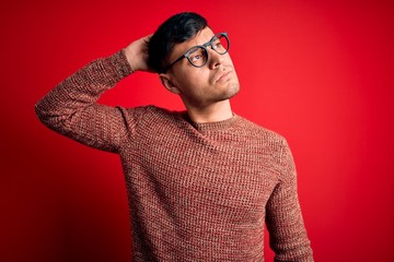 Young handsome hispanic man wearing nerd glasses over red background confuse and wondering about...