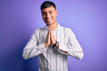 Young handsome hispanic man wearing elegant business shirt standing over purple background praying with hands together asking for forgiveness smiling confident.