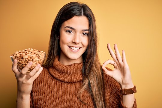 Young beautiful girl holding bowl of salty peanuts standing over yellow background doing ok sign with fingers, excellent symbol