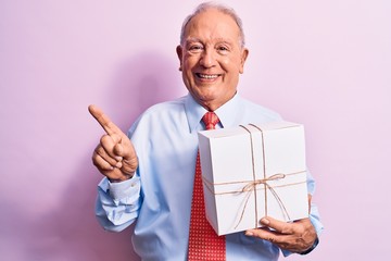 Senior grey-haired businessman wearing tie holding birthday gift over pink background smiling happy pointing with hand and finger to the side
