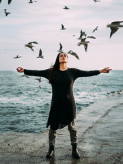 A young girl in a black dress and jeans stands by the sea with a strong wind. Hair and seagulls fly. The power of thought, magic, self-awareness, gaining strength to overcome difficulties. Peace