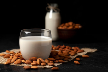 almond milk without sugar and lactose in glass with sprinkled nuts, vegetarian milk drink on a dark black table