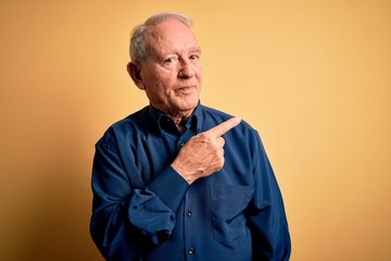 Grey haired senior man wearing casual blue shirt standing over yellow background Pointing with hand finger to the side showing advertisement, serious and calm face