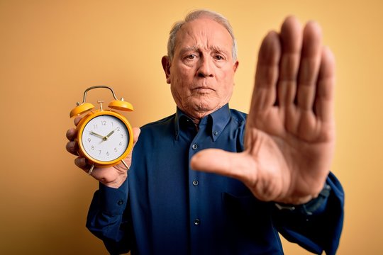 Senior grey haired man holding vintage alarm clock over yellow background with open hand doing stop sign with serious and confident expression, defense gesture