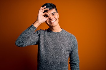 Young handsome man wearing casual sweater standing over isolated orange background doing ok gesture with hand smiling, eye looking through fingers with happy face.