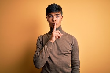 Young handsome man wearing casual turtleneck sweater over isolated yellow background asking to be quiet with finger on lips. Silence and secret concept.