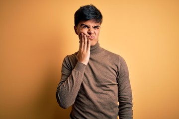 Young handsome man wearing casual turtleneck sweater over isolated yellow background touching mouth with hand with painful expression because of toothache or dental illness on teeth. Dentist