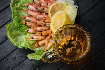 Boiled prawns on a black plate and a glass of beer on a wooden table. Shrimp with lemon on a lettuce leaf. Beer in a glass mug