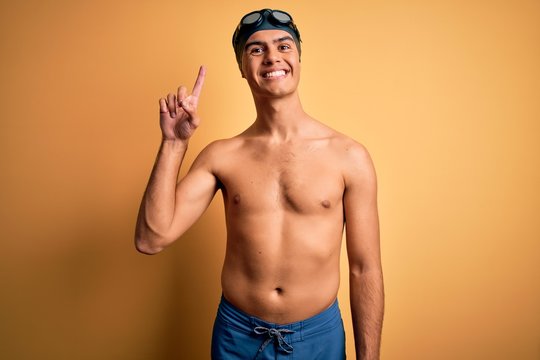 Young handsome man shirtless wearing swimsuit and swim cap over isolated yellow background showing and pointing up with finger number one while smiling confident and happy.