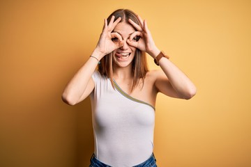 Young beautiful redhead woman wearing casual t-shirt over isolated yellow background doing ok gesture like binoculars sticking tongue out, eyes looking through fingers. Crazy expression.