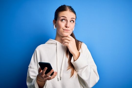 Young redhead sportswoman doing sport listening to music using earphones and smartphone with hand on chin thinking about question, pensive expression. Smiling with thoughtful face. Doubt concept.