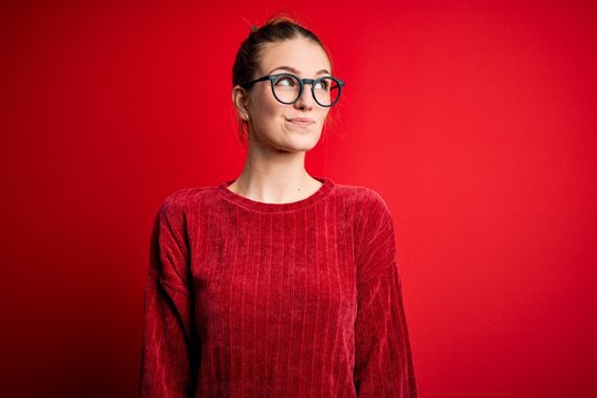 Young beautiful redhead woman wearing casual sweater over isolated red background smiling looking to the side and staring away thinking.