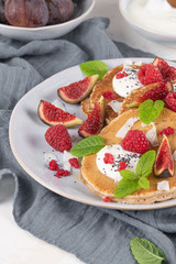 Pancakes with raspberries, figs, yogurt, coconut zest, honey and mint leaves on a plate.