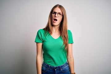 Obraz premium Young beautiful redhead woman wearing casual green t-shirt and glasses over white background In shock face, looking skeptical and sarcastic, surprised with open mouth