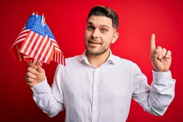 Young man with blue eyes holding flag of united states of america over red isolated background surprised with an idea or question pointing finger with happy face, number one