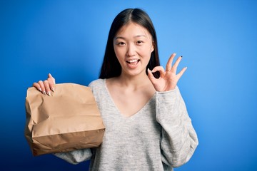 Young asian woman holding delivery paper bag for takeaway food over blue isolated background doing ok sign with fingers, excellent symbol