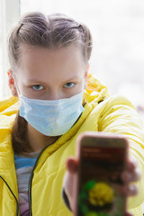 Girl at home with phone near window in isolation in medical mask.