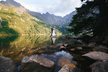 The bride and groom are standing on a stone in the lake hugging each other on a background of mountains. Wide angle