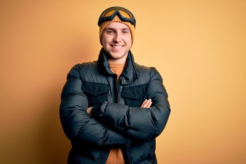 Young handsome caucasian man wearing hat, coat and ski glasses for winter and snow weather happy face smiling with crossed arms looking at the camera. Positive person.