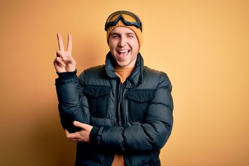 Young handsome caucasian man wearing hat, coat and ski glasses for winter and snow weather smiling with happy face winking at the camera doing victory sign. Number two.
