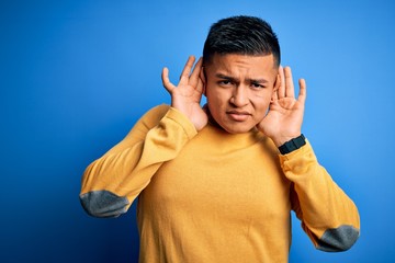 Young handsome latin man wearing yellow casual sweater over isolated blue background Trying to hear both hands on ear gesture, curious for gossip. Hearing problem, deaf