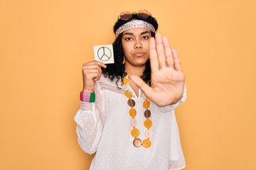 African american curly hippie woman wearing sunglasses holding reminder with peace symbol with open hand doing stop sign with serious and confident expression, defense gesture