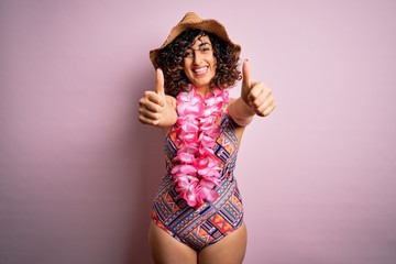 Young beautiful arab woman on vacation wearing swimsuit and hawaiian lei flowers approving doing positive gesture with hand, thumbs up smiling and happy for success. Winner gesture.