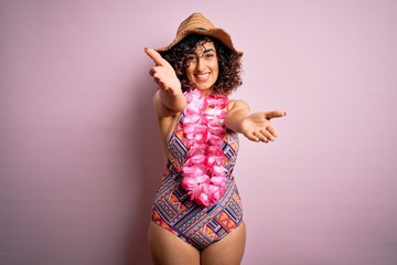 Young beautiful arab woman on vacation wearing swimsuit and hawaiian lei flowers looking at the camera smiling with open arms for hug. Cheerful expression embracing happiness.