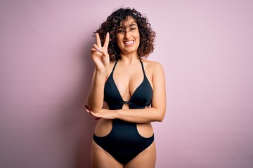 Obraz premium Young beautiful arab woman on vacation wearing swimsuit and sunglasses over pink background smiling with happy face winking at the camera doing victory sign. Number two.