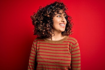 Young beautiful curly arab woman wearing casual striped sweater standing over red background looking away to side with smile on face, natural expression. Laughing confident.