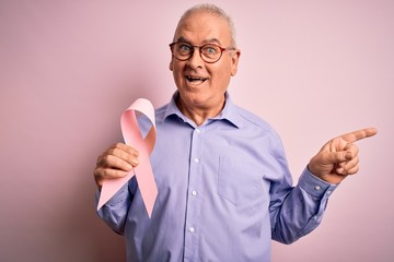 Middle age hoary man holding pink cancer ribbon symbol over isolated background very happy pointing with hand and finger to the side