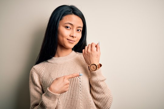 Young beautiful chinese woman wearing casual sweater over isolated white background In hurry pointing to watch time, impatience, looking at the camera with relaxed expression