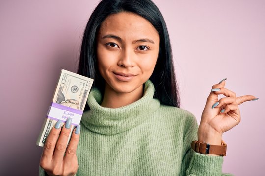 Young beautiful chinese woman holding dollars standing over isolated pink background very happy pointing with hand and finger to the side