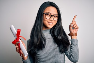 Young beautiful chinese woman holding diploma standing over isolated white background surprised...