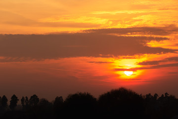 Bright glow of the sun disc through the clouds during sunset in the countryside above the forest and tree silhouettes. Scenic night landscape for background or poster. Weather change