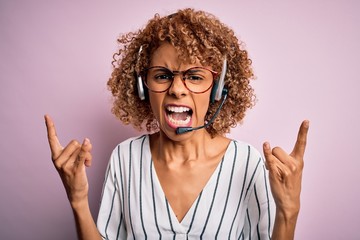 African american curly call center agent woman working using headset over pink background shouting with crazy expression doing rock symbol with hands up. Music star. Heavy concept.