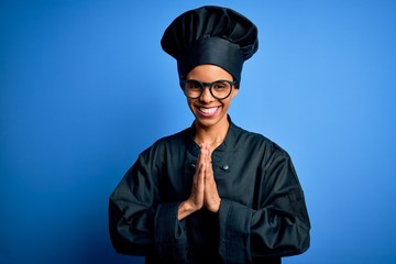 Young african american chef woman wearing cooker uniform and hat over blue background praying with hands together asking for forgiveness smiling confident.