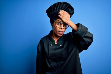 Young african american chef woman wearing cooker uniform and hat over blue background surprised...