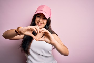 Young brunette woman wearing casual sport cap over pink background smiling in love doing heart symbol shape with hands. Romantic concept.