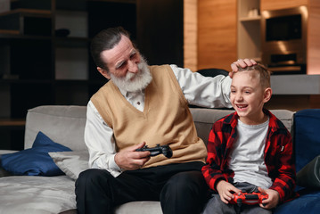 Fototapeta premium Cute little boy with grandfather sitting on sofa and playing video game with game pad