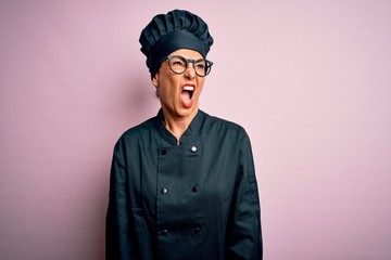 Middle age brunette chef woman wearing cooker uniform and hat over isolated pink background angry and mad screaming frustrated and furious, shouting with anger. Rage and aggressive concept.