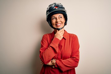 Middle age motorcyclist woman wearing motorcycle helmet over isolated white background with hand on chin thinking about question, pensive expression. Smiling with thoughtful face. Doubt concept.