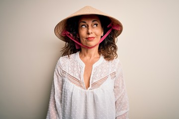 Middle age brunette woman wearing asian traditional conical hat over white background smiling looking to the side and staring away thinking.