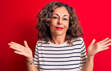 Middle age beautiful brunette woman wearing striped t-shirt standing over red background clueless and confused with open arms, no idea and doubtful face.