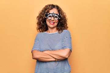 Middle age beautiful woman controlling vision using optometry glasses over yellow background happy...