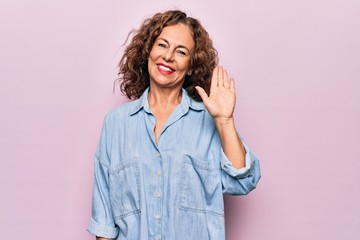 Middle age beautiful woman wearing casual denim shirt standing over pink background Waiving saying hello happy and smiling, friendly welcome gesture